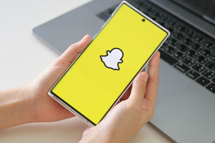 Smartphone showing Snapchat logo held by hands with laptop in the background, related to ultimatums and lost friendships.