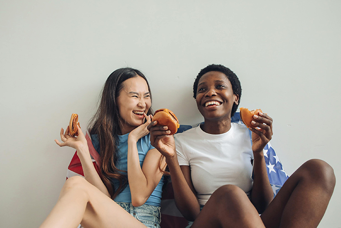 Two women sitting with an American flag, smiling and eating burgers, capturing a casual and friendly moment. Two women sitting with an American flag, smiling and eating burgers, capturing a casual and friendly moment.