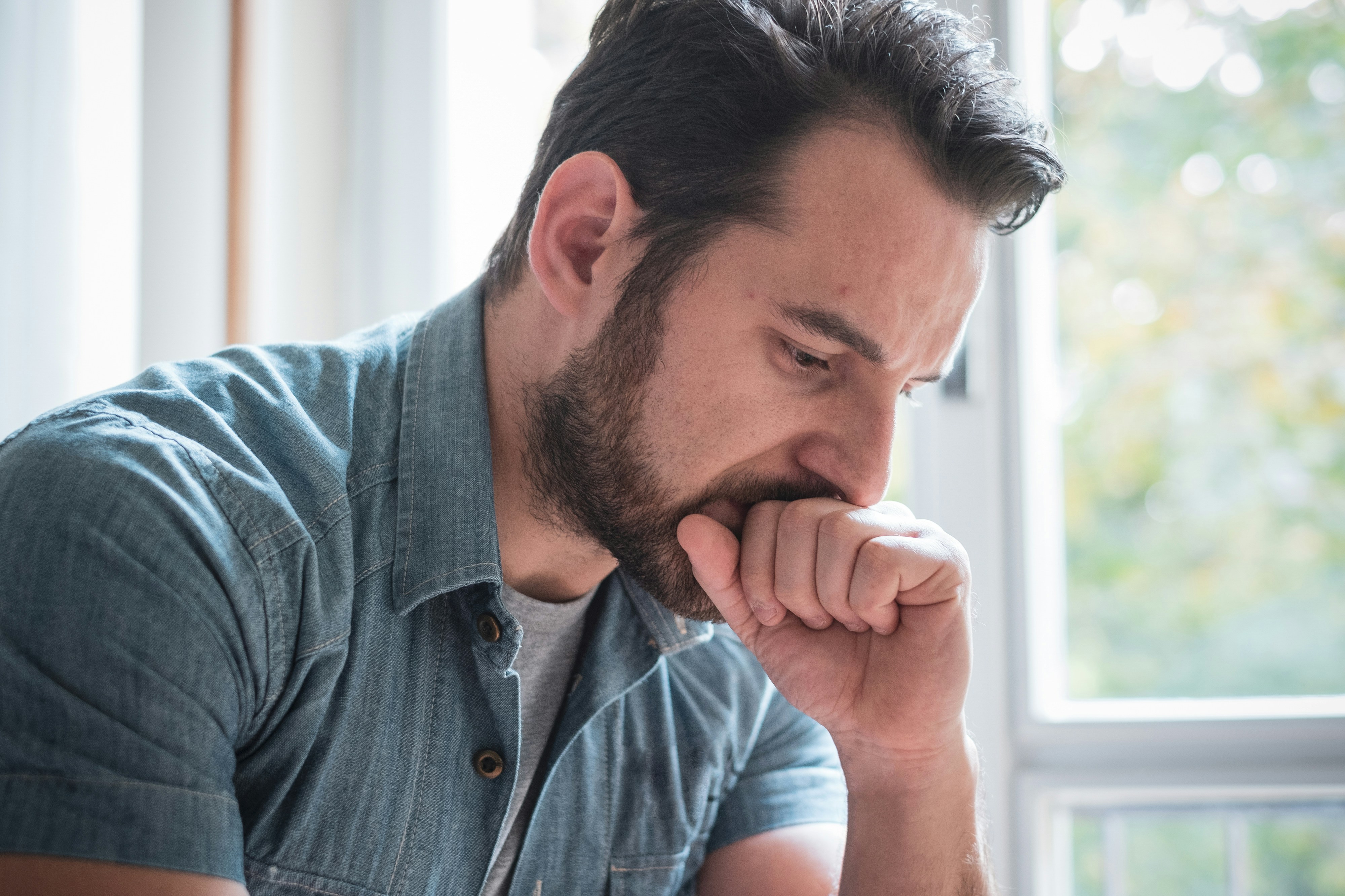 Man in denim shirt looking distressed while sitting near a window symbolizing childhood best friend favor conflict and humiliation. Man in denim shirt looking distressed while sitting near a window symbolizing childhood best friend favor conflict and humiliation.