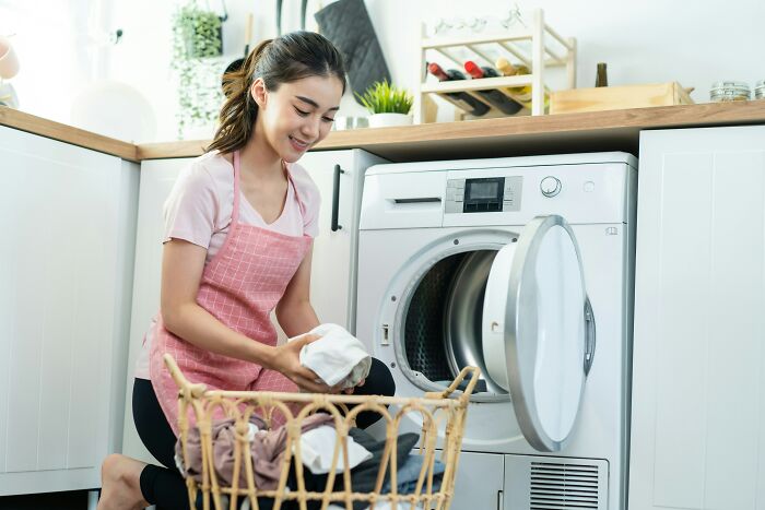 Young woman kneeling by washing machine, holding laundry with a smile, showing a tiny thing that made trip better.
