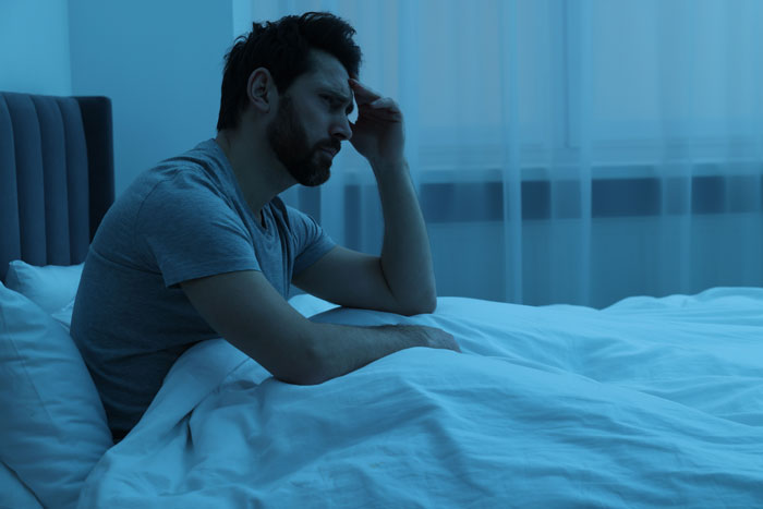 Man sitting on bed in bedroom at night, appearing worried as he questions future with girlfriend over son's demands.