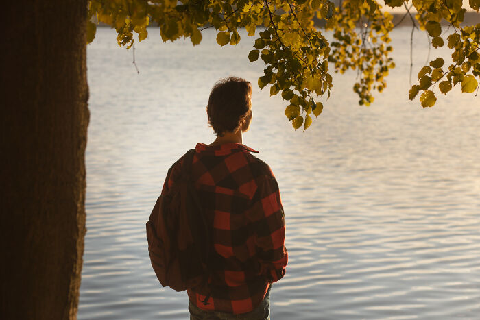 Man wearing red plaid shirt and backpack standing by a lake, reflecting on life after losing an amazing partner.