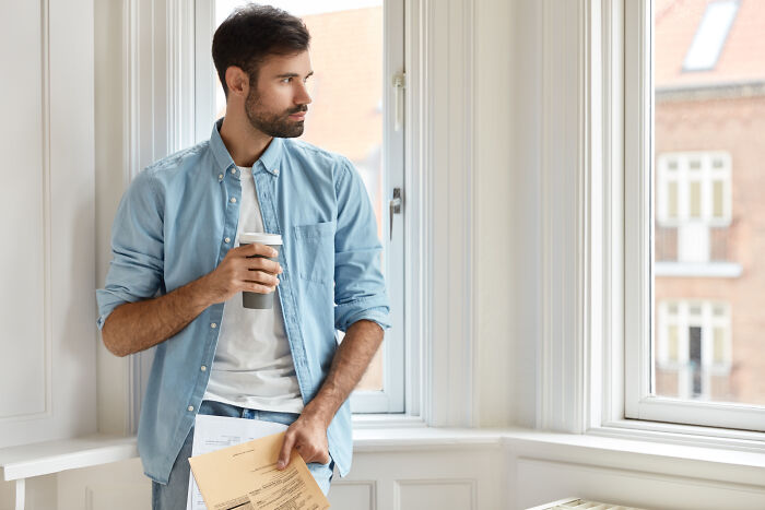Man reflecting by a window holding papers and a coffee cup, representing men who lost amazing partners and their life changes.