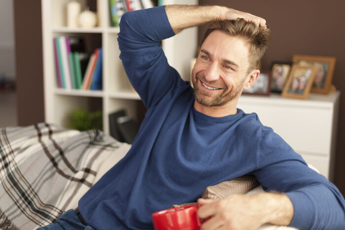 Man smiling thoughtfully on a couch holding a red mug, representing men who lost amazing partners and their new life challenges.