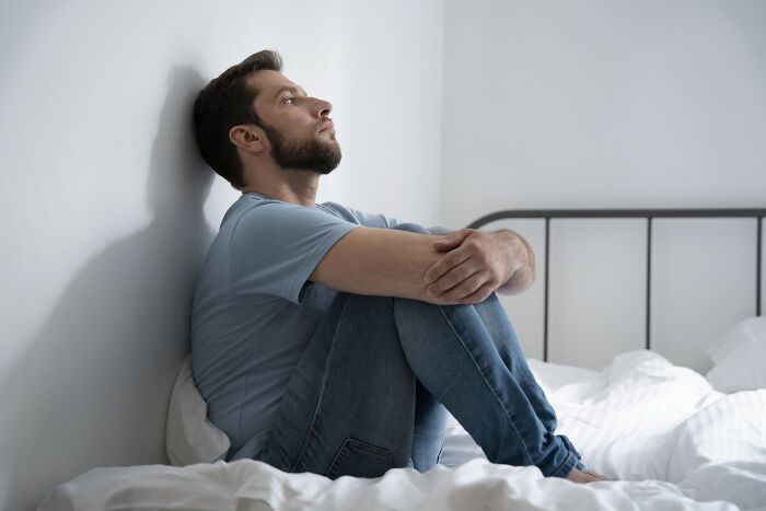 Man sitting on bed, hugging knees and looking thoughtful, reflecting on life after losing an amazing partner.