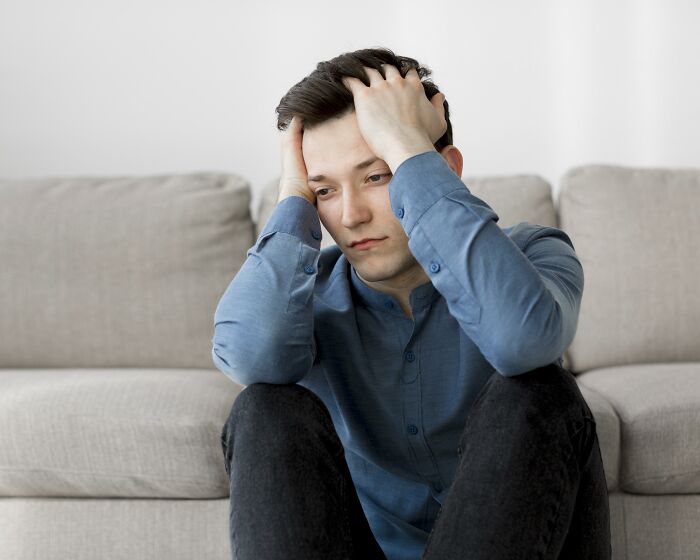 Young man in blue shirt sitting on floor, holding head in hands, reflecting on life after losing amazing partner.