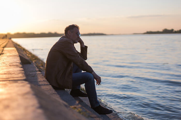 Man sitting alone by the water at sunset, reflecting on life after losing an amazing partner and coping with changed realities.