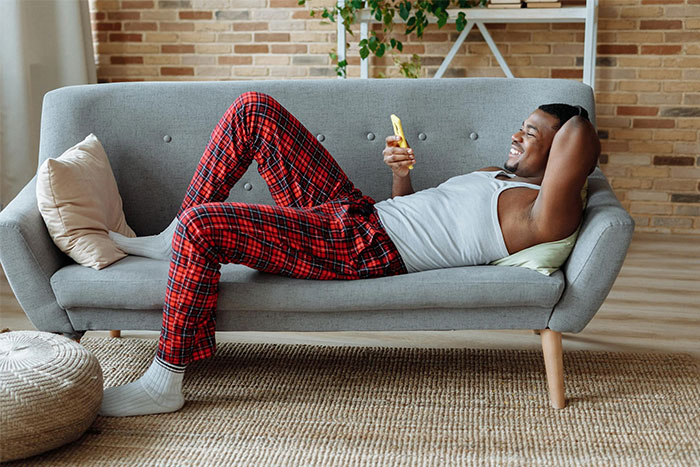 Man in red plaid pants relaxing on a gray sofa, smiling while looking at his phone in a cozy living room setting. Man in red plaid pants relaxing on a gray sofa, smiling while looking at his phone in a cozy living room setting.