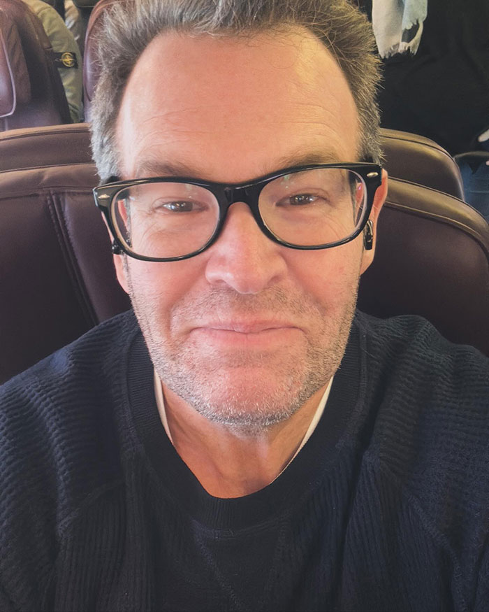 Man with glasses and stubble wearing a black sweater, seated and smiling gently in a close-up selfie. Man with glasses and stubble wearing a black sweater, seated and smiling gently in a close-up selfie.
