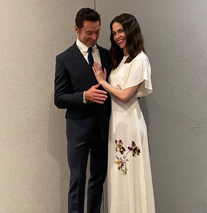 Hugh Jackman and Sutton Foster smiling together, dressed formally against a plain gray wall background. Hugh Jackman and Sutton Foster smiling together, dressed formally against a plain gray wall background.