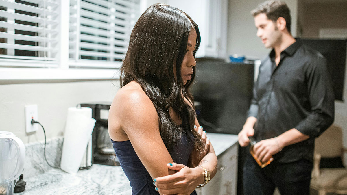Woman with crossed arms looks upset in kitchen while husband stands behind holding a bottle, reflecting conflict after cross-country move.