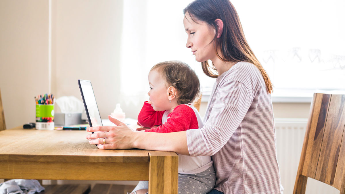 Mother working on a tablet while holding her toddler, highlighting challenges of refusal to help with child care.