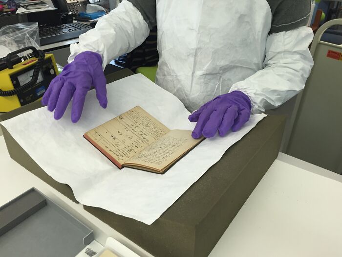 Person wearing protective gear and purple gloves handling an old notebook related to Marie Curie research archives.