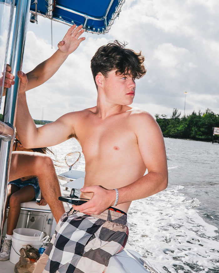 Shirtless Jack Doherty on a boat holding a phone, enjoying a sunny day on the water with scenic greenery in the background.