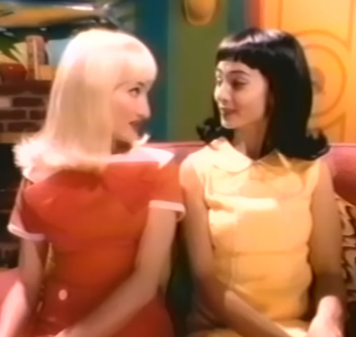 Two women sitting closely on a couch, one with blonde hair and the other with black hair, in a retro setting. Two women sitting closely on a couch, one with blonde hair and the other with black hair, in a retro setting.