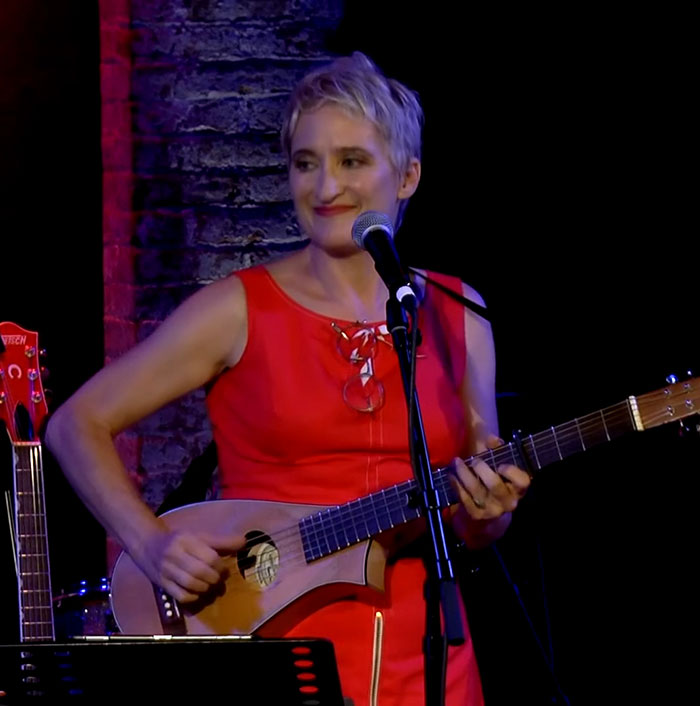 Singer Jill Sobule performing onstage in a red dress playing guitar and singing into a microphone at a live event. Singer Jill Sobule performing onstage in a red dress playing guitar and singing into a microphone at a live event.