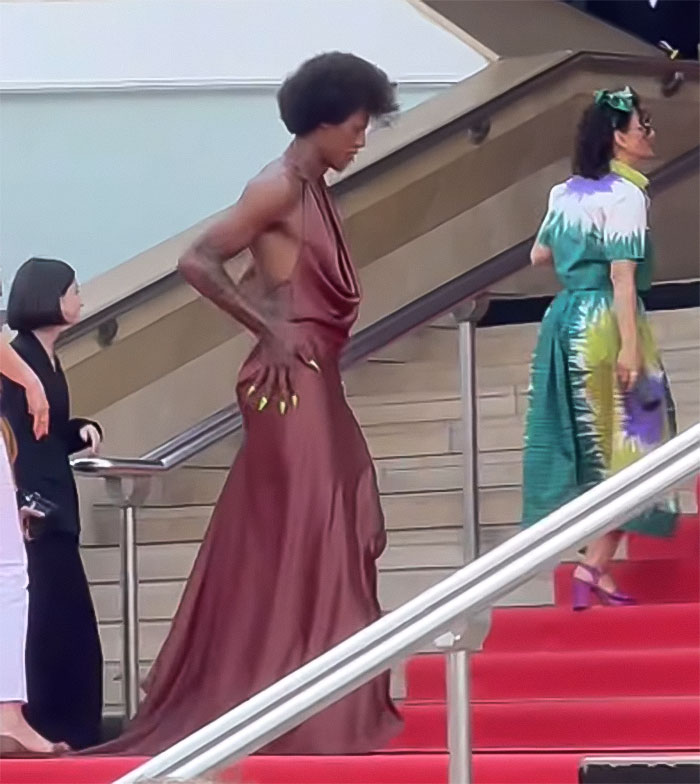 Star in a flowing brown dress revealing backside, breaking Cannes dress code while walking up red carpet stairs. Star in a flowing brown dress revealing backside, breaking Cannes dress code while walking up red carpet stairs.