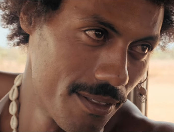 Close-up of a man with a mustache and curly hair wearing a shell necklace, highlighting natural facial features and expression. Close-up of a man with a mustache and curly hair wearing a shell necklace, highlighting natural facial features and expression.