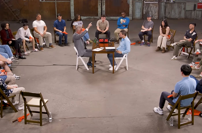 Jordan Peterson seated at a table debating atheists in a large room with an audience watching closely. Jordan Peterson seated at a table debating atheists in a large room with an audience watching closely.