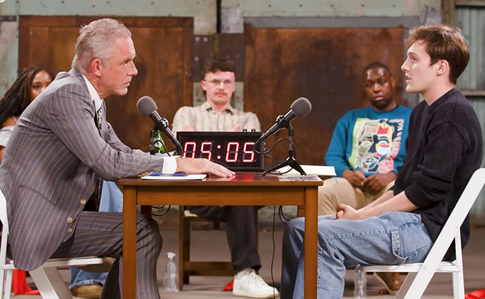 Jordan Peterson in a formal debate setting, facing a young atheist opponent, with microphones and a digital timer on the table. Jordan Peterson in a formal debate setting, facing a young atheist opponent, with microphones and a digital timer on the table.