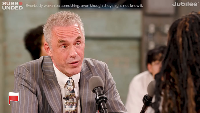 Jordan Peterson in a debate setting, wearing a striped suit, discussing ideas during a controversial atheist dialogue. Jordan Peterson in a debate setting, wearing a striped suit, discussing ideas during a controversial atheist dialogue.