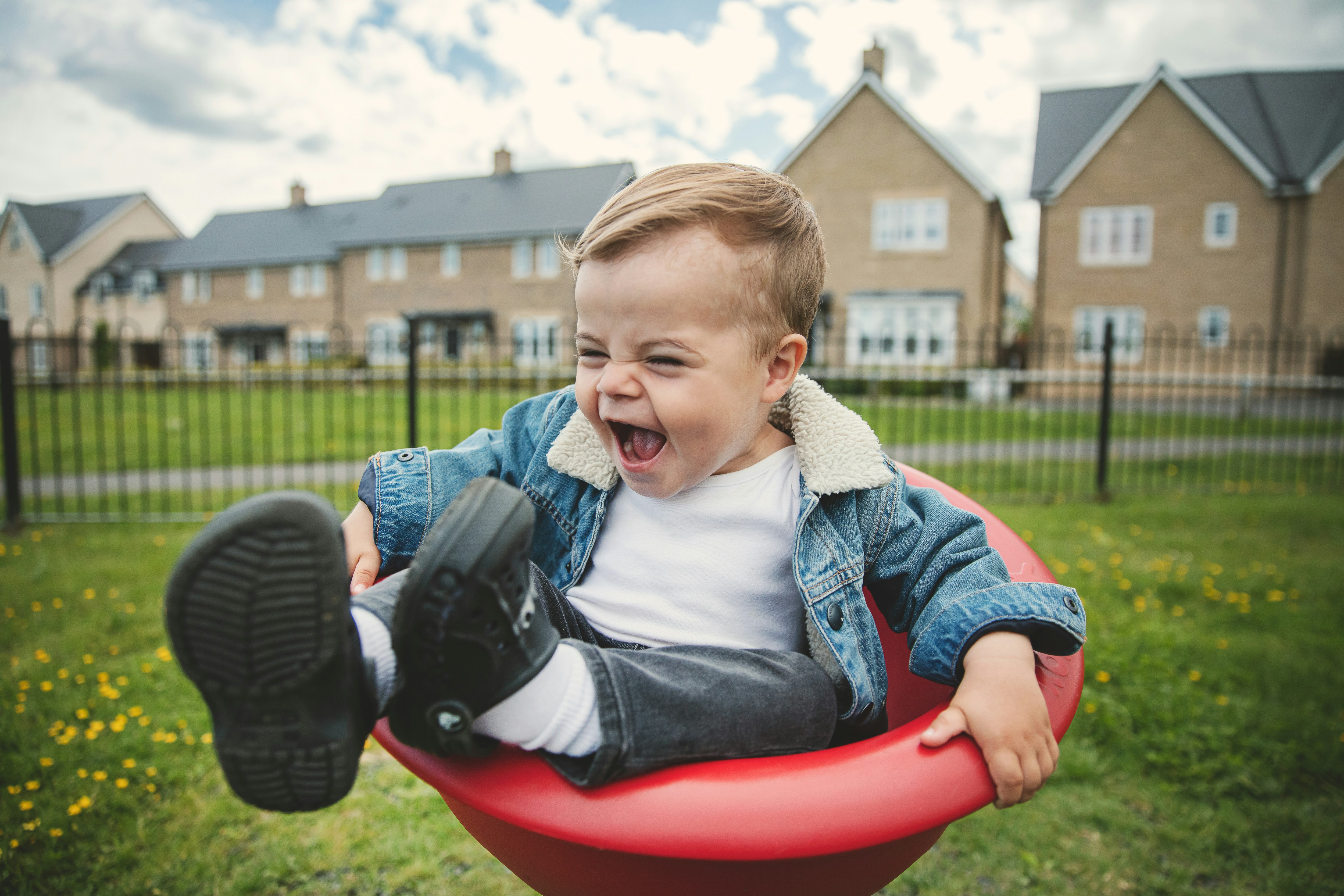 Toddler laughing joyfully outdoors in a red swing, capturing playful moments with major side-eye from parents nearby. Toddler laughing joyfully outdoors in a red swing, capturing playful moments with major side-eye from parents nearby.
