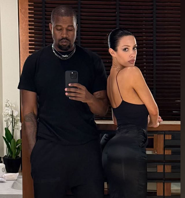Bianca Censori posing in black outfit next to a man taking a mirror selfie in a room with wooden blinds. Bianca Censori posing in black outfit next to a man taking a mirror selfie in a room with wooden blinds.