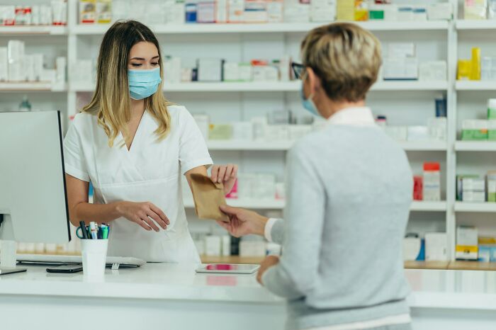 Pharmacist wearing a mask handing a paper bag to a customer in a pharmacy with shelves of medicine behind.
