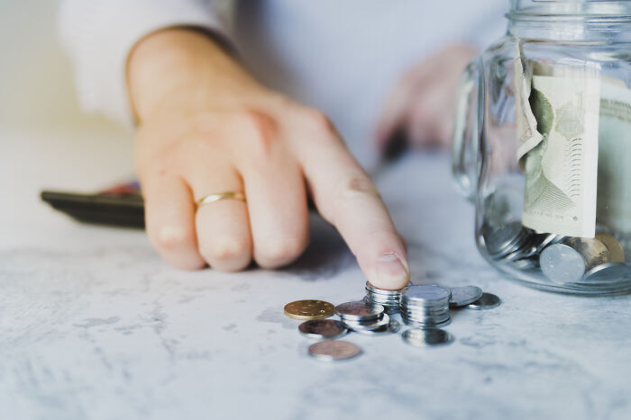 Person with a gold ring stacking coins and counting money next to a jar of cash, reflecting unhinged parents diabolical behavior concept.