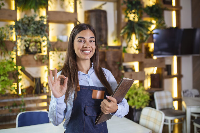 Smiling woman in apron holding menu and making OK gesture in a cozy cafe, representing Karen unhinged parents diabolical.