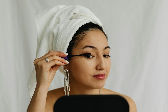 Woman with towel on head applying mascara in a mirror, revealing industry secrets behind closed doors in beauty routines.