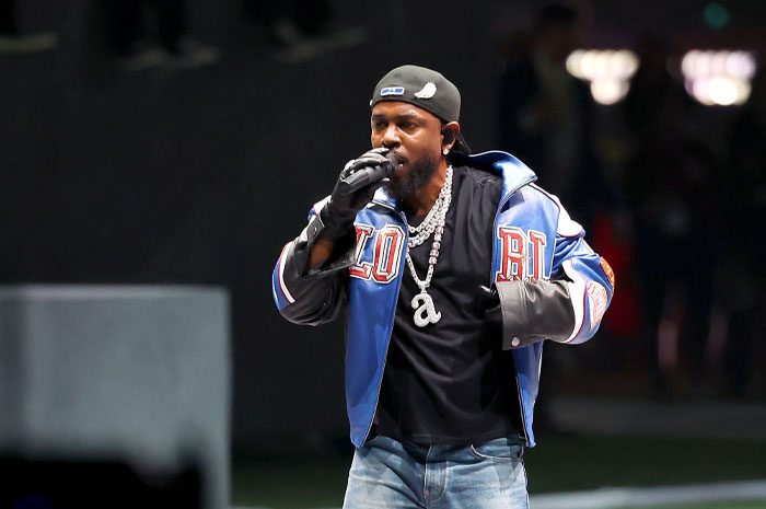 Kendrick Lamar performing on stage wearing a blue jacket and chains, showcasing his rise in net worth and success.