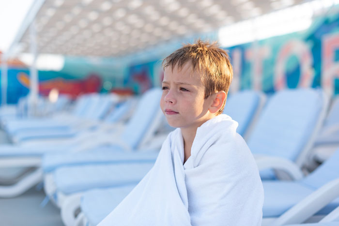 Young boy wrapped in a towel sitting alone on a poolside lounge chair at a condo pool area on a sunny day