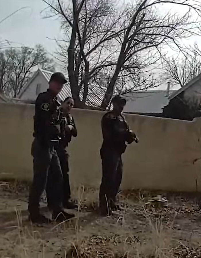 Three police officers standing outside near a fence in a grassy area during daytime in a residential neighborhood. Three police officers standing outside near a fence in a grassy area during daytime in a residential neighborhood.