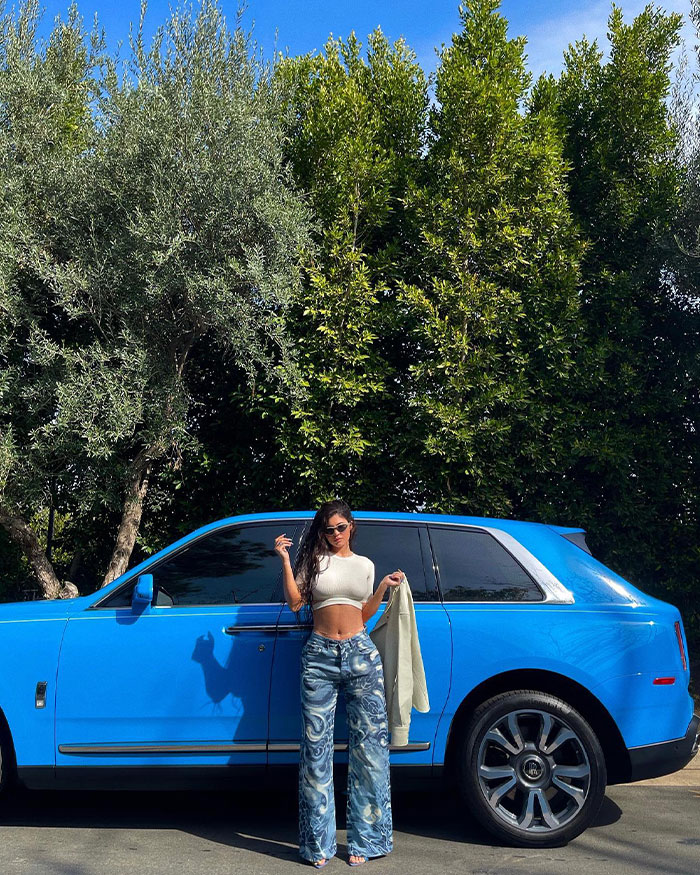 Kylie Junner posing by a blue luxury car surrounded by trees, highlighting Kylie Jenner’s net worth and financial status update.