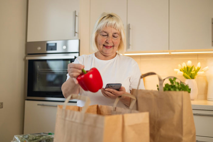 Woman holding red bell pepper and phone while unpacking groceries in kitchen, related to grocery Karen shopping dispute. Woman holding red bell pepper and phone while unpacking groceries in kitchen, related to grocery Karen shopping dispute.