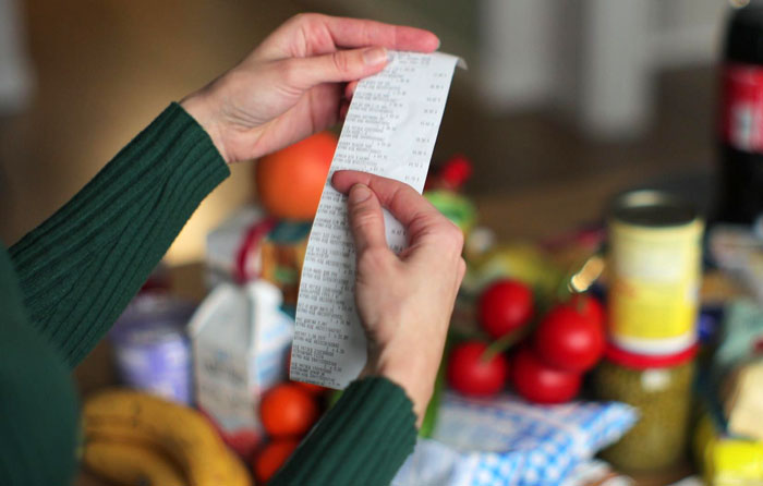 Person holding a grocery receipt with various fresh produce and packaged food items on a table nearby. Person holding a grocery receipt with various fresh produce and packaged food items on a table nearby.