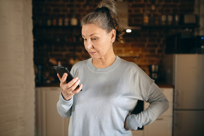 Middle-aged woman looking at smartphone with concern in kitchen, representing grocery Karen refusing to pay for extra items. Middle-aged woman looking at smartphone with concern in kitchen, representing grocery Karen refusing to pay for extra items.