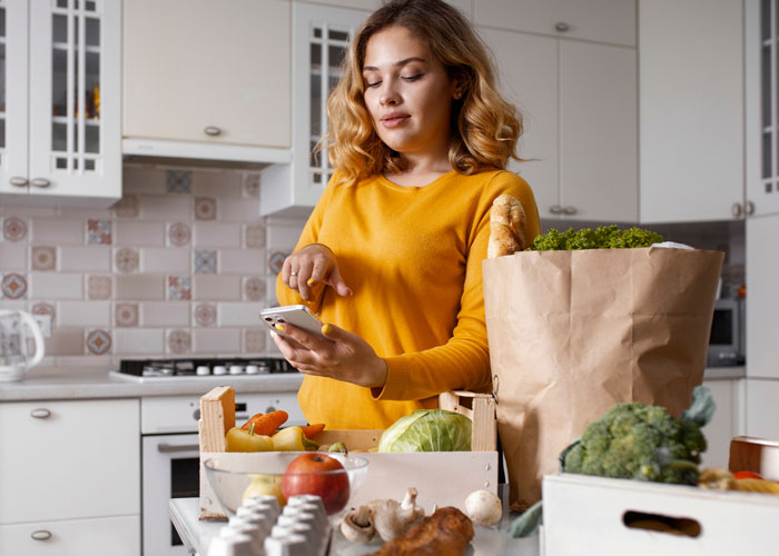 Woman in kitchen with groceries, using phone, illustrating grocery Karen refusing to pay for extra items scenario. Woman in kitchen with groceries, using phone, illustrating grocery Karen refusing to pay for extra items scenario.