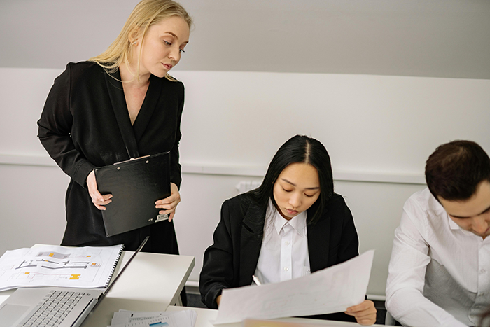 A boss in black blazer reviews work while a young employee focuses on documents, showing a lazy employee learning to work. A boss in black blazer reviews work while a young employee focuses on documents, showing a lazy employee learning to work.