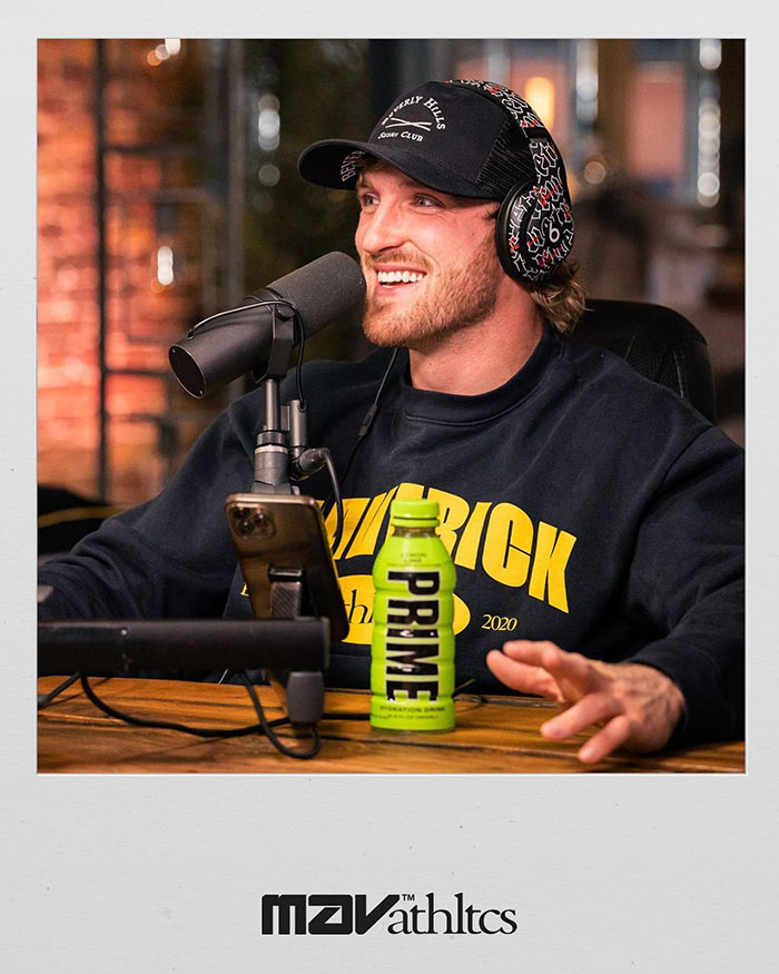 Logan Paul speaking into a microphone wearing headphones and a cap with a Prime drink bottle on the table.
