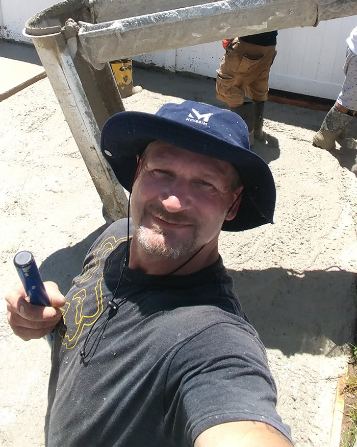Man wearing a blue hat and gray shirt taking a selfie at a construction site with cement and workers in the background Man wearing a blue hat and gray shirt taking a selfie at a construction site with cement and workers in the background