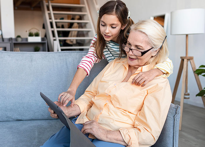 Young girl with grandmother on sofa using a tablet, illustrating mom furious over MIL's weird gift to pregnancy-obsessed child. Young girl with grandmother on sofa using a tablet, illustrating mom furious over MIL's weird gift to pregnancy-obsessed child.
