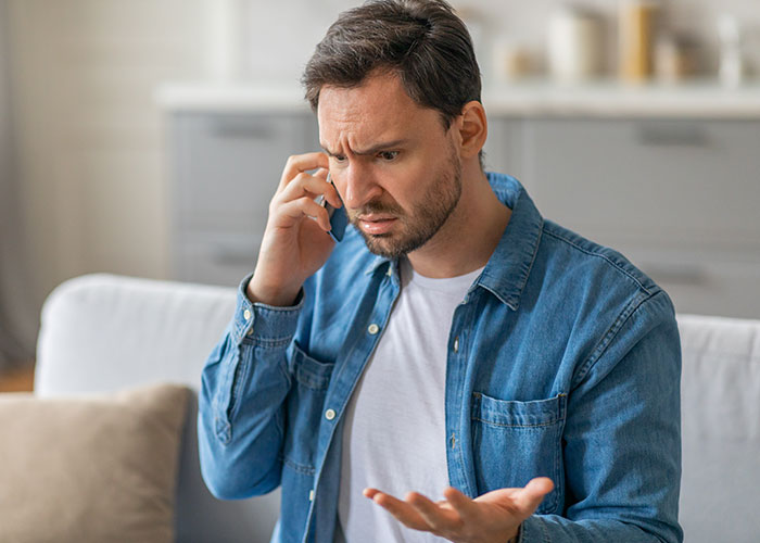 Man in a denim shirt on phone looking confused and frustrated while sitting on a couch in a modern living room. Man in a denim shirt on phone looking confused and frustrated while sitting on a couch in a modern living room.