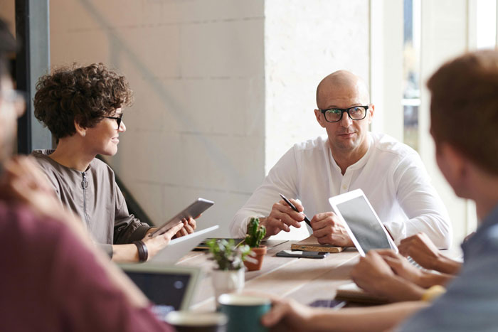 Manager discussing employee concerns with team, holding pen, with others using tablets in a modern office meeting room. Manager discussing employee concerns with team, holding pen, with others using tablets in a modern office meeting room.