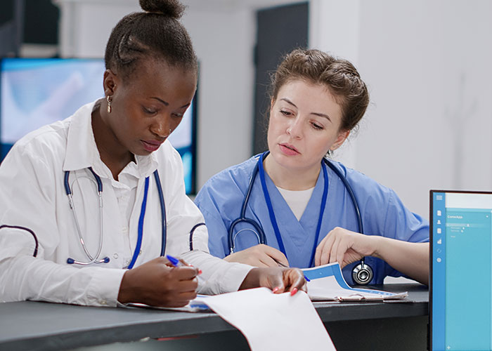 Two medical workers in scrubs and lab coats reviewing documents together in a busy medical facility. Two medical workers in scrubs and lab coats reviewing documents together in a busy medical facility.