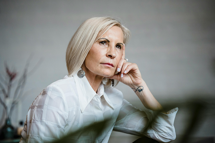 Older woman with blonde hair wearing white shirt and silver earrings, looking concerned while resting her head on her hand. Older woman with blonde hair wearing white shirt and silver earrings, looking concerned while resting her head on her hand.