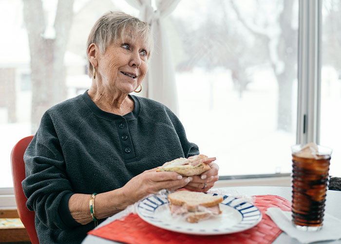 Elderly woman sitting by the window eating fast food sandwich with a drink, highlighting mom refuses to leave kids with MIL. Elderly woman sitting by the window eating fast food sandwich with a drink, highlighting mom refuses to leave kids with MIL.