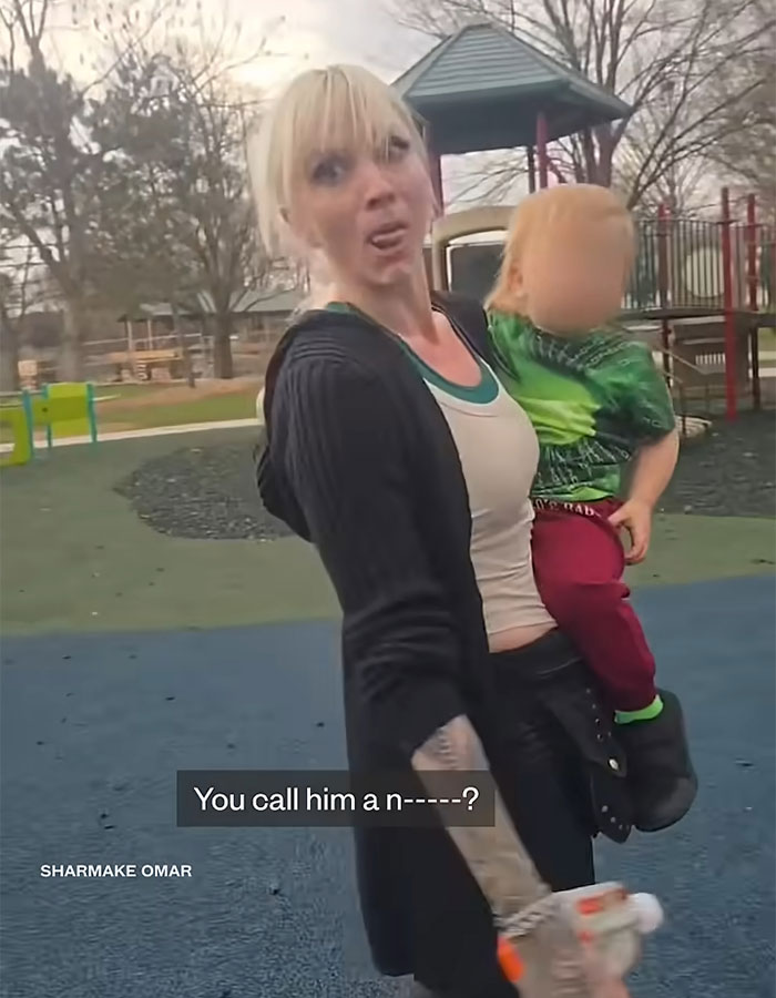 Woman accused of racist slur on playground holding child with blurred face in a park setting during confrontation. Woman accused of racist slur on playground holding child with blurred face in a park setting during confrontation.