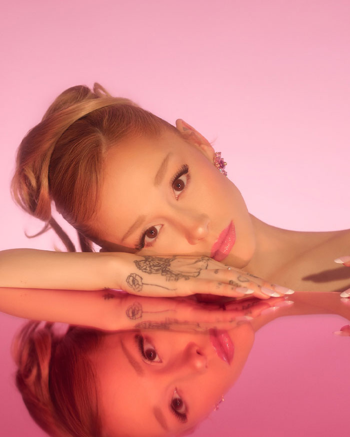 Ariana Grande with tattoos resting her face on a reflective surface, exploring the science behind most beautiful woman.